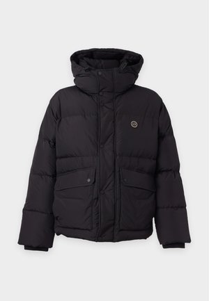 Homme portant une casquette beige, une veste matelassée noire, un polo bleu marine, un pantalon beige et des bottines noires à lacets, debout devant un fond uni.