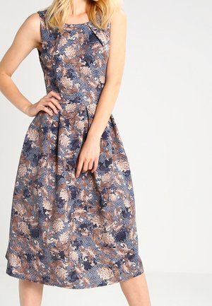 Frau trägt ein ärmelloses knielanges Kleid mit blauem, braunem und beigem Blumenmuster, steht mit einer Hand auf der Hüfte.