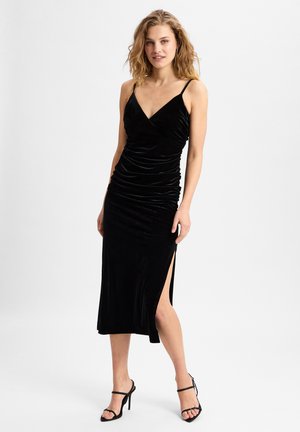 Schwarzes Samt-Midikleid mit V-Ausschnitt, gerafftem Design, seitlichem Schlitz und dünnen Schulterträgern. Das Model trägt schwarze High-Heels.