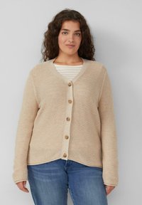 Beige, gebreide cardigan met een V-hals, lange mouwen en vijf houten knopen aan de voorkant. Guti over een wit gestreept shirt.