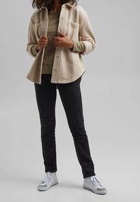 Beige Hemdjacke mit zwei Brusttaschen, über einem grünen Oberteil getragen, kombiniert mit schwarzen Hosen und weißen Sneakers mit grauen Akzenten.