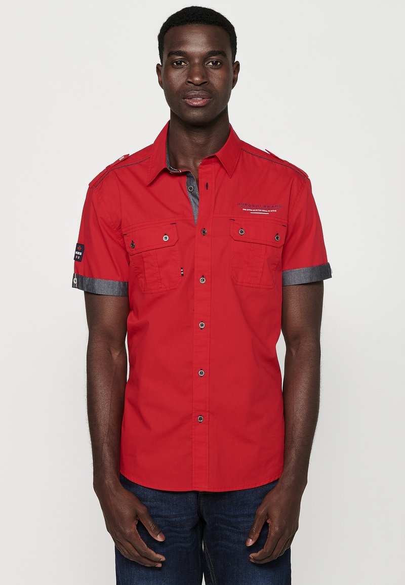 Camisa roja de manga corta con un cuello gris, dos bolsillos en el pecho y detalles de botones. La tela parece suave; cuenta con puños de manga en contraste.