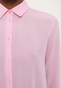 Blouse longue à manches transparentes rose clair avec un col classique, dotée de boutons assortis sur le devant pour la fermeture. Texture lisse et délicate.