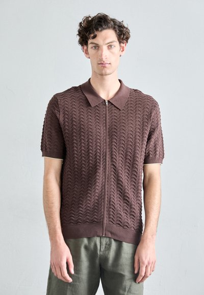 Polo a maniche corte marrone con un motivo a onde testurizzato, dotato di zip frontale e colletto a contrasto, abbinato a pantaloni verde chiaro.