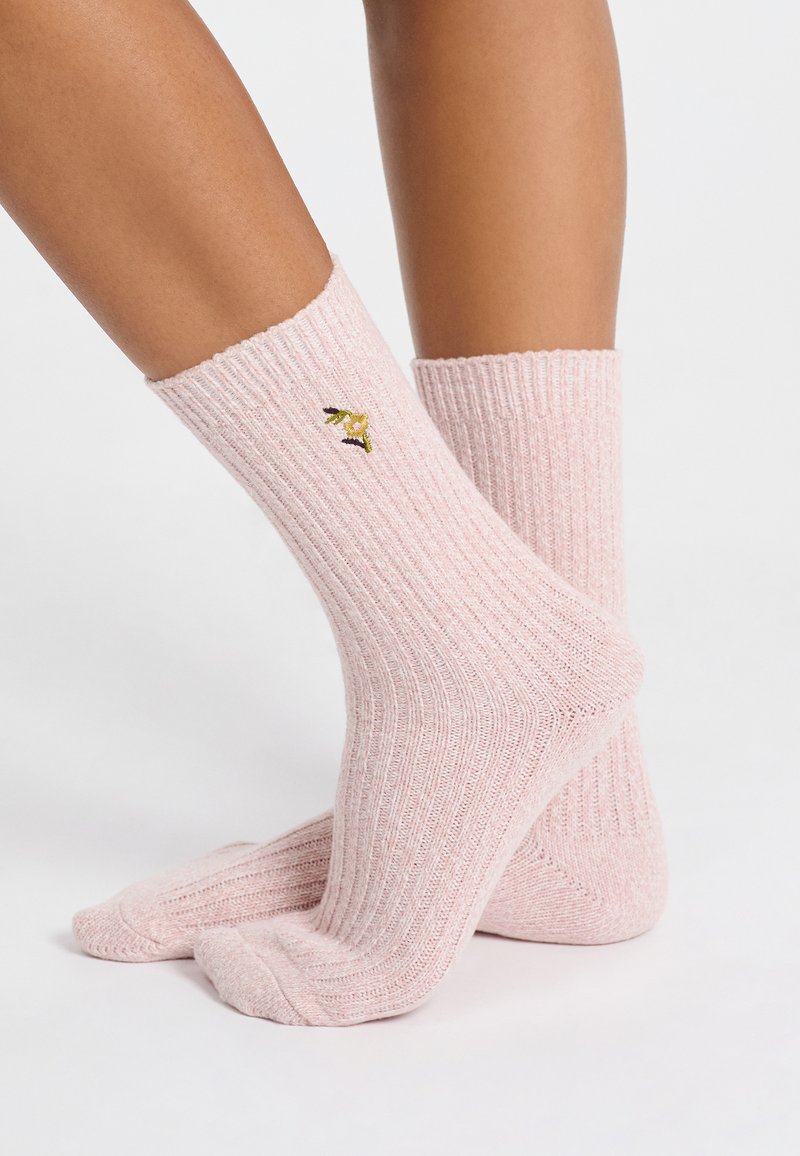 Piernas que llevan calcetines de caña corta de rayas rosa con una pequeña flor amarilla bordada y hojas verde oscuro en el costado.