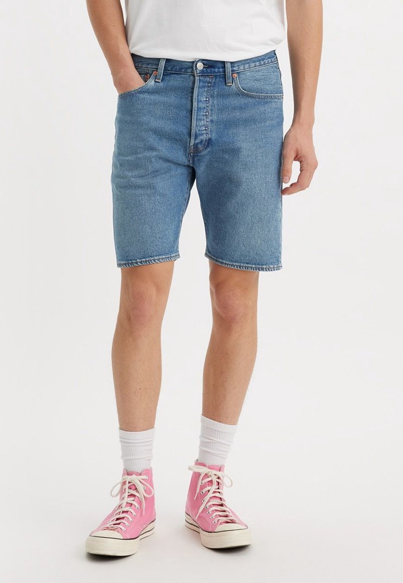 Homme portant un short en jean bleu, des chaussettes blanches et des baskets montantes roses, debout avec une main dans la poche sur un fond uni.