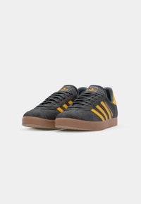 Black suede sneakers with yellow stripes, rubber sole, and textured detailing. Features embroidered logo on tongue and side.