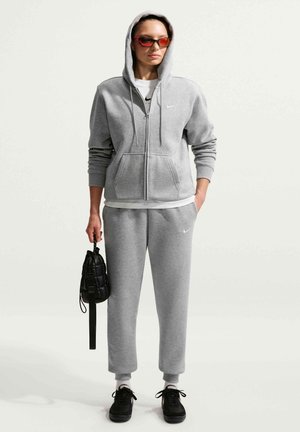 Kobieta w szarej bluzie z kapturem i dresie, czarne sportowe buty i czerwone okulary przeciwsłoneczne, trzyma małą czarną torbę jedną ręką.