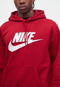 Άτομο φοράει κόκκινο φούτερ Nike με λευκό λογότυπο και ορατά κορδόνια, εμφανίζεται από τον λαιμό μέχρι τη μέση πάνω σε απλό φόντο.