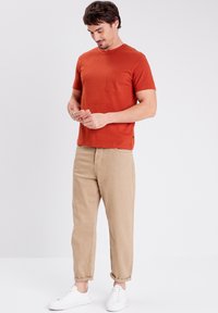 T-shirt a maniche corte color ruggine con taschino quadrato, abbinato a pantaloni beige chiaro arrotolati e sneakers bianche. Design semplice con collo a giro.