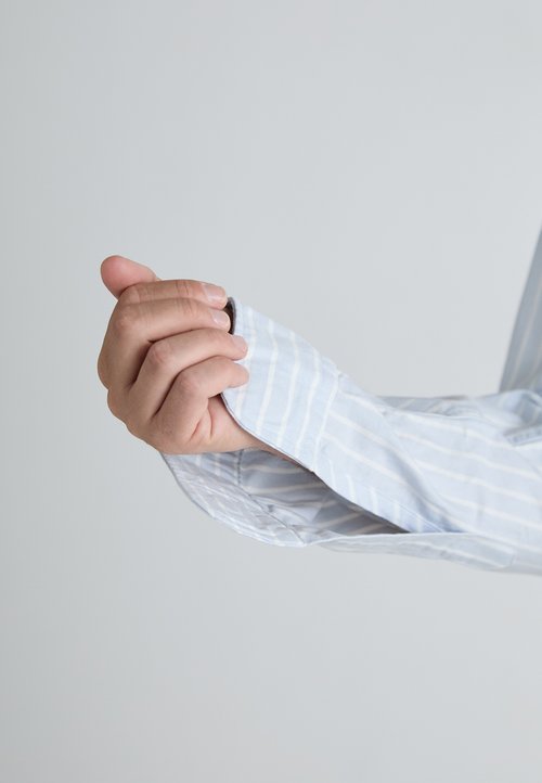 Chemise bleu clair à col avec rayures blanches, présentant des poignets retroussés, une texture de tissu lisse, et un détail de bouton visible sur la manche.