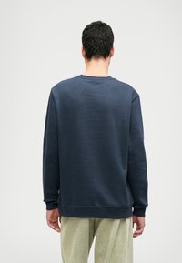 Felpa blu navy con maniche lunghe, collo rotondo e tessuto testurizzato. Presenta polsini e orlo a costine. Indossata con pantaloni di colore chiaro.