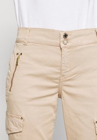 Béžové cargo kalhoty s kapsou na zip na stehně, dvěma knoflíkovými zapínáními v pase a jemnou, hladkou texturou.