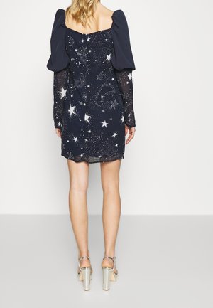 Femme portant une mini-robe bleu marine avec des épaules bouffantes et un imprimé céleste étoilé, associée à des sandales à talons hauts argentées, debout de dos.