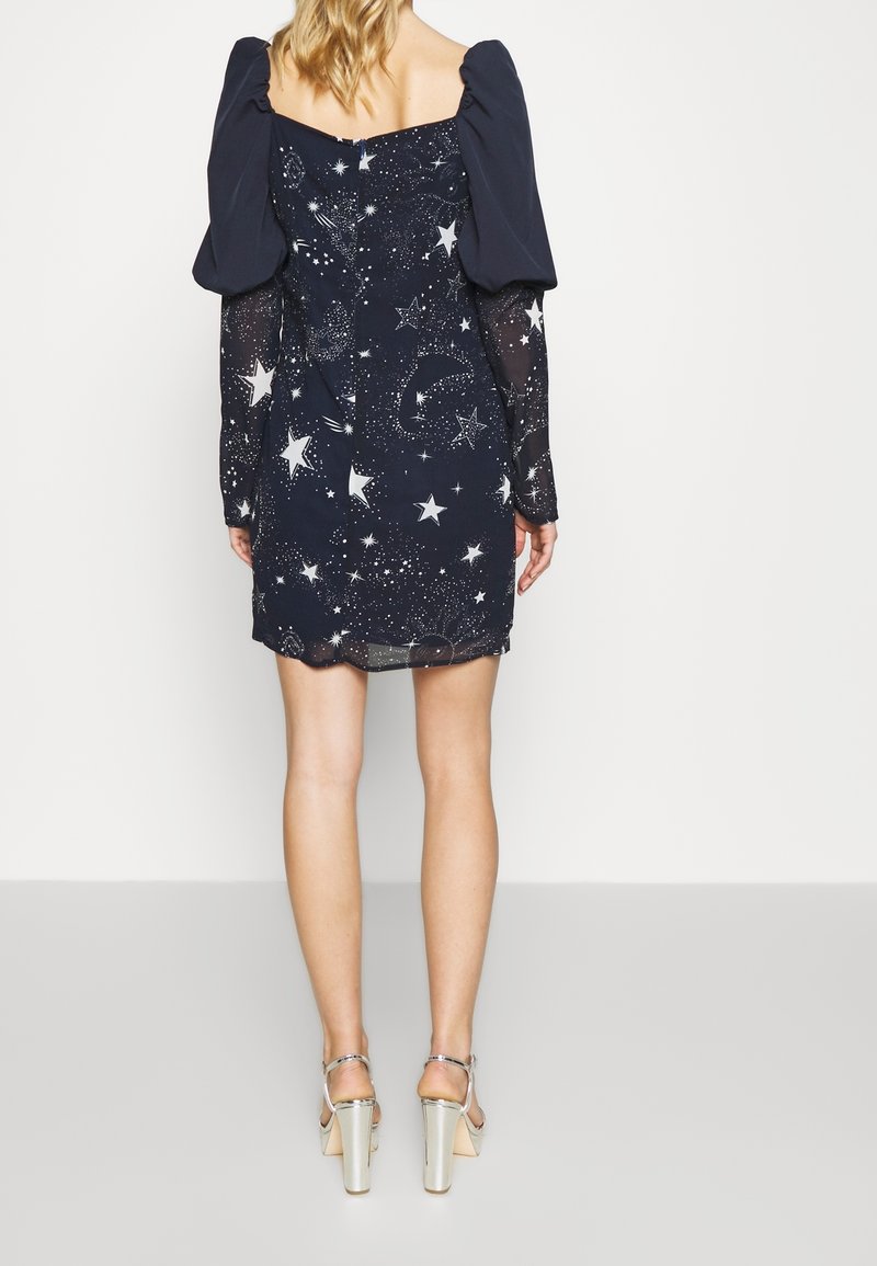Femme portant une mini-robe bleu marine avec des épaules bouffantes et un imprimé céleste étoilé, associée à des sandales à talons hauts argentées, debout de dos.