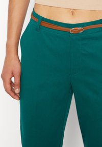 Tyrkysové kalhoty s hnědým koženým pásem a stříbrnou sponou. Hladký materiál s dobře padnoucím střihem a jemnými švy.