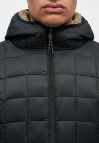 Giubbotto trapuntato nero con chiusura zip e cappuccio foderato in tessuto beige. La superficie è liscia con una finitura opaca.