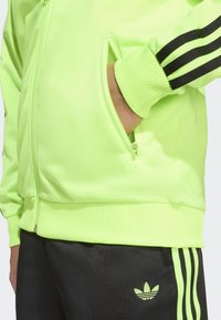 Neongrön jacka med dragkedja, ribbade muddar och kant, med svarta tre-randiga detaljer på ärmar och sidoficka. Svarta byxor med logotyp.