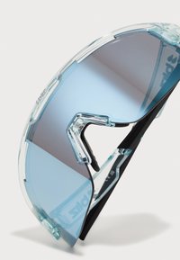 Lunettes de soleil avec une monture transparente bleu clair et des verres gris dégradés. Présentent un design incurvé et des accents noirs linéaires sur les branches.