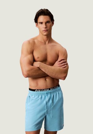 Homme musclé debout, bras croisés, portant un short de bain bleu clair avec une ceinture noire Calvin Klein visible sur un fond uni.