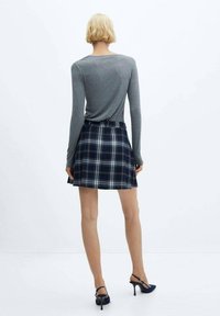 Haut à manches longues gris, jupe à carreaux marine avec des accents blancs et verts, et chaussures à talons noires. La texture semble lisse et le design est décontracté.