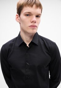 Zwarte langemouwen shirt gemaakt van een gladde stof, heeft een klassieke kraag en een knoopsluiting aan de voorkant met zichtbare tonale knopen, aansluitende pasvorm.