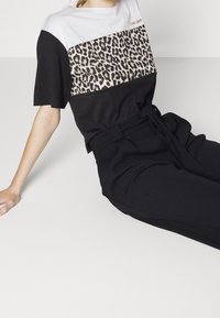 Mujer vestida con una blusa de manga corta en negro, blanco y estampado de leopardo, combinada con pantalones negros de tiro alto con un cinturón anudado, sentada sobre una superficie blanca.