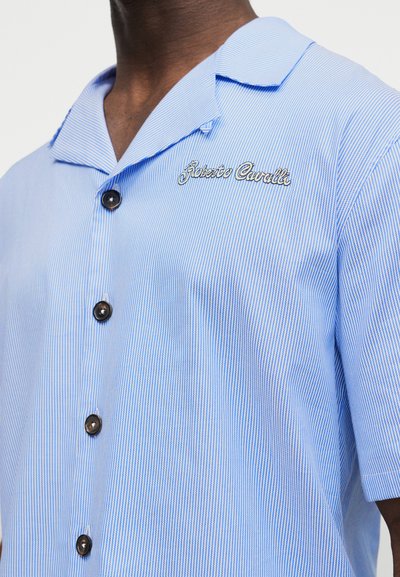 Άνδρας που φοράει ανοιφτό μπλε, λεπτή ριγέ κοντομάνικη πουκάμισο με σκούρα κουμπιά και κεντημένο κείμενο "Roberto Cavalli" στο στήθος.