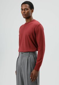 Rode trui met lange mouwen van zacht materiaal, gecombineerd met grijze, taillebroek met dubbele voorplooien en knopdetails.