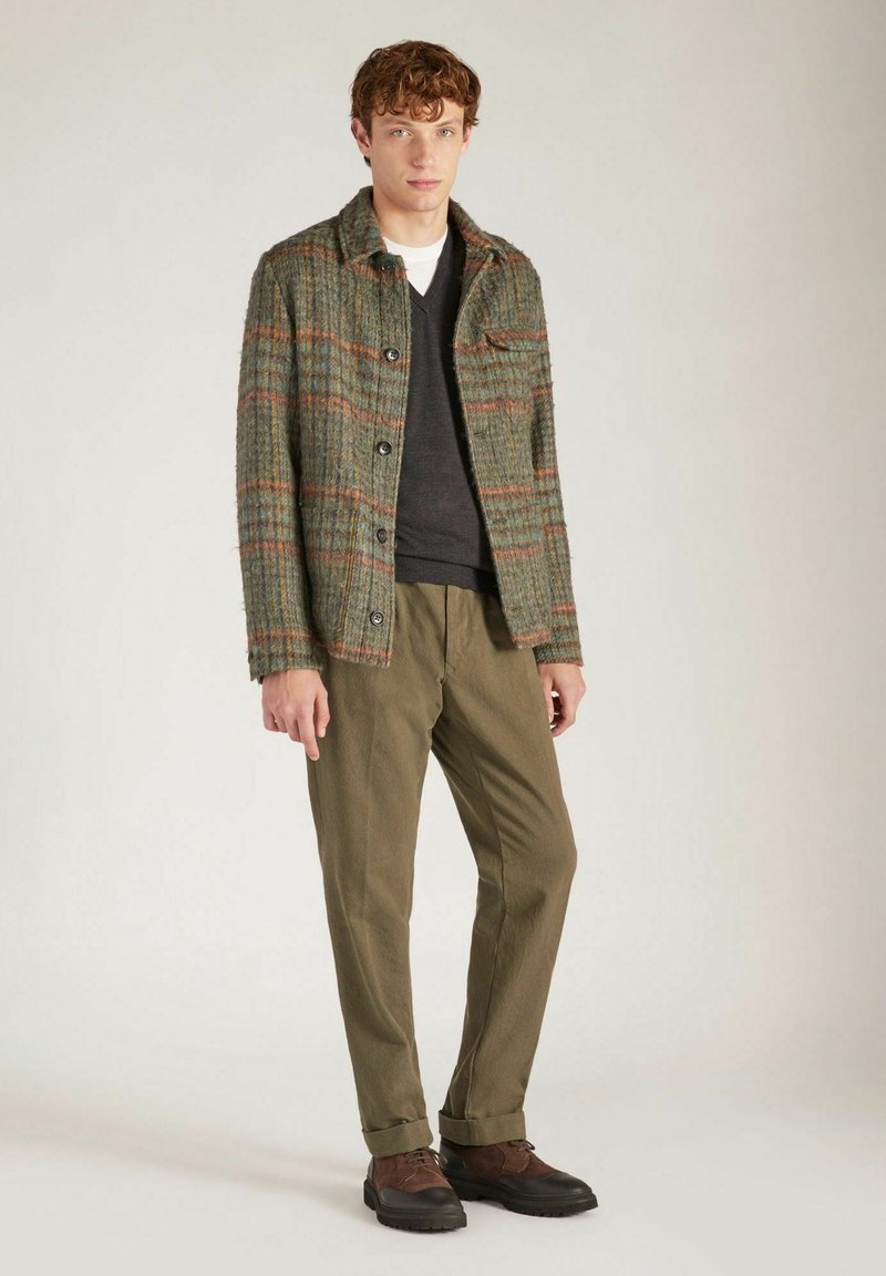 Veste en mélange de laine verte avec un motif à carreaux orange, fermeture à boutons, poche poitrine. Associée à un pantalon olive et des chaussures à lacets marron.