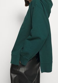 Teal hoodie med avslappnad passform, ribbade ärmslut och en känguruficka. Den har mjukt bomullstyg och sidoslitsar för ökad rörelsefrihet.