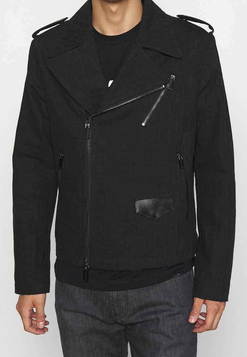 Man med en svart motorcykeljacka med asymmetriska silverdragkedjor, skulderklappar och en liten läderfickficka på framsidan.