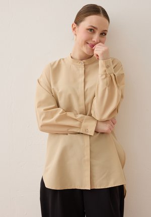 Ung kvinde med lys hud og håret samlet bagud smiler, iført en beige skjorte med knapper og sorte bukser, står mod en ensfarvet væg.