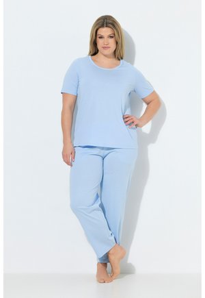 Vrouw staat op blote voeten tegen een witte muur, gekleed in een lichtblauw shirt met korte mouwen en bijpassende losse broek.