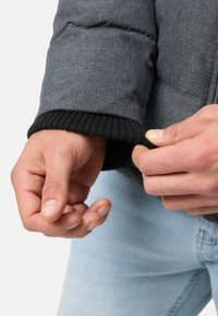 Szary pikowany płaszcz z prążkowanym czarnym mankietem, trzymany w dłoni. Tkanina ma teksturowane wykończenie, a mankiet ściśle przylega do nadgarstka.