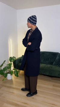 Hosszú tengerészkék kabát kockás ing felett, barna bő szárú nadrág, fekete cipő és mintás sapka. Zöld kanapé és növény a háttérben.