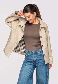 Femme aux cheveux bruns en queue de cheval portant un manteau beige boutonné par-dessus une chemise marron et un jean bleu, ajustant le col du manteau et regardant vers le bas.