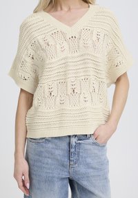 Világos bézs kötött pulóver V-nyakkal, rövid ujjakkal és bonyolult nyílt mintákkal, csíkos díszítéssel, farmernadrággal párosítva.