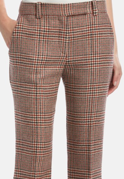 Woman wearing high-waist pants with red, beige, and black plaid pattern, hands in pockets, against a white background.