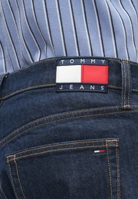 Κοντινό πλάνο από σκούρο μπλε τζιν παντελόνι με λογότυπο Tommy Jeans και ριγέ ανοιχτό μπλε πουκάμισο στο εσωτερικό.