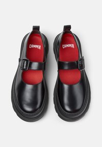 Zwarte Mary Jane-schoenen met dikke zolen, gespbanden en rode binnenzolen met het merk "Camper", gezien van bovenaf op een witte achtergrond.