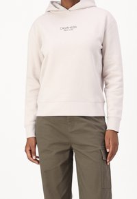 Sudadera con capucha de color beige claro, con un corte relajado y puños de canalé, que presenta un pequeño logo centrado en gris "Calvin Klein New York".