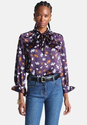 Femme portant un chemisier en satin violet à motif floral avec un nœud au cou, un jean bleu, des boucles d'oreilles créoles dorées et des bracelets dorés à son poignet gauche.