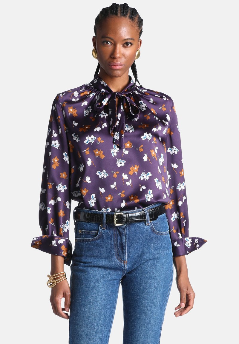 Femme portant un chemisier en satin violet à motif floral avec un nœud au cou, un jean bleu, des boucles d'oreilles créoles dorées et des bracelets dorés à son poignet gauche.
