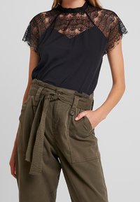 Top nero con maniche a cappuccio in pizzo e scollatura alta, abbinato a pantaloni verdi oliva con cintura, caratterizzati da una finitura testurizzata e tasche.