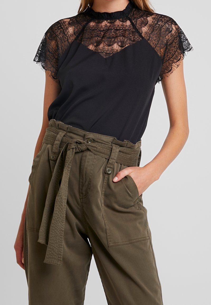 Top nero con maniche a cappuccio in pizzo e scollatura alta, abbinato a pantaloni verdi oliva con cintura, caratterizzati da una finitura testurizzata e tasche.