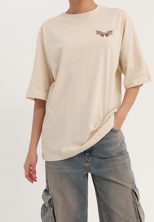 Person iført beige oversized T-shirt med "Sleepy Club" vinge-logo på brystet og falmede blå cargobukser med hånden i lommen.