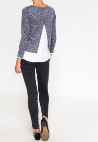 Marinblått texturerat tröja med öppning på ryggen, bärs över en vit topp och svarta jeans i skinny-fit. Outfiten avslutas med svarta lägenheter.