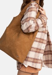 Hnědá kožená kabelka přes rameno s dvěma vnějšími kapsami, hladkým povrchem a minimalistickým designem, nošena přes rameno na pozadí kostkované bundy.