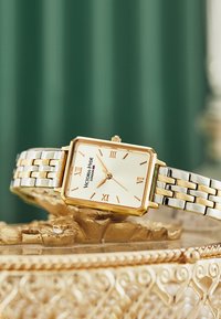 Reloj rectangular chapado en oro con una esfera color crema, números romanos y una pulsera metálica de dos tonos, colocado sobre una superficie decorativa.
